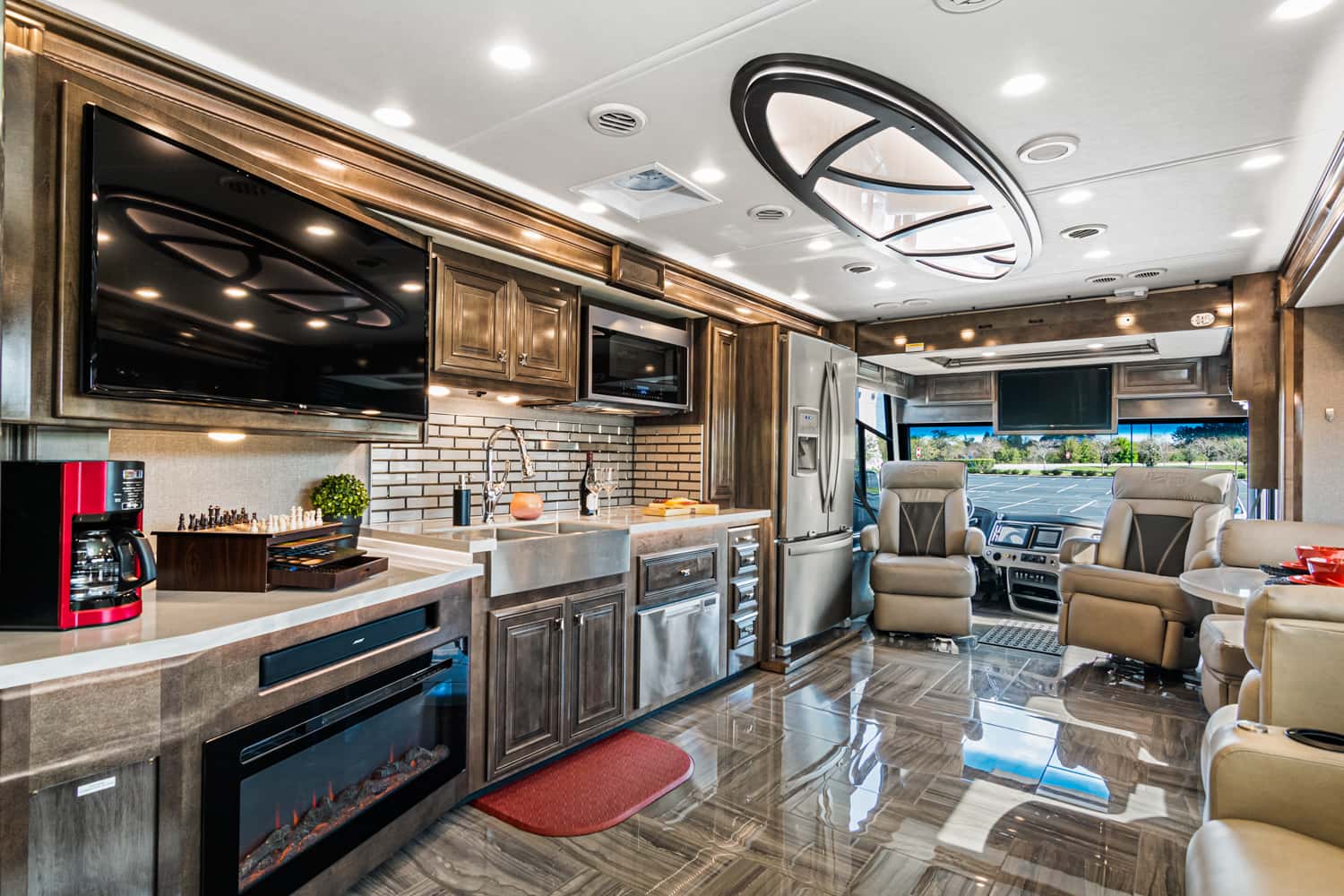 Interior of a modern RV with kitchen and lounge area, featuring a large TV, stainless steel appliances, leather seating, a fireplace, and tiled flooring.
