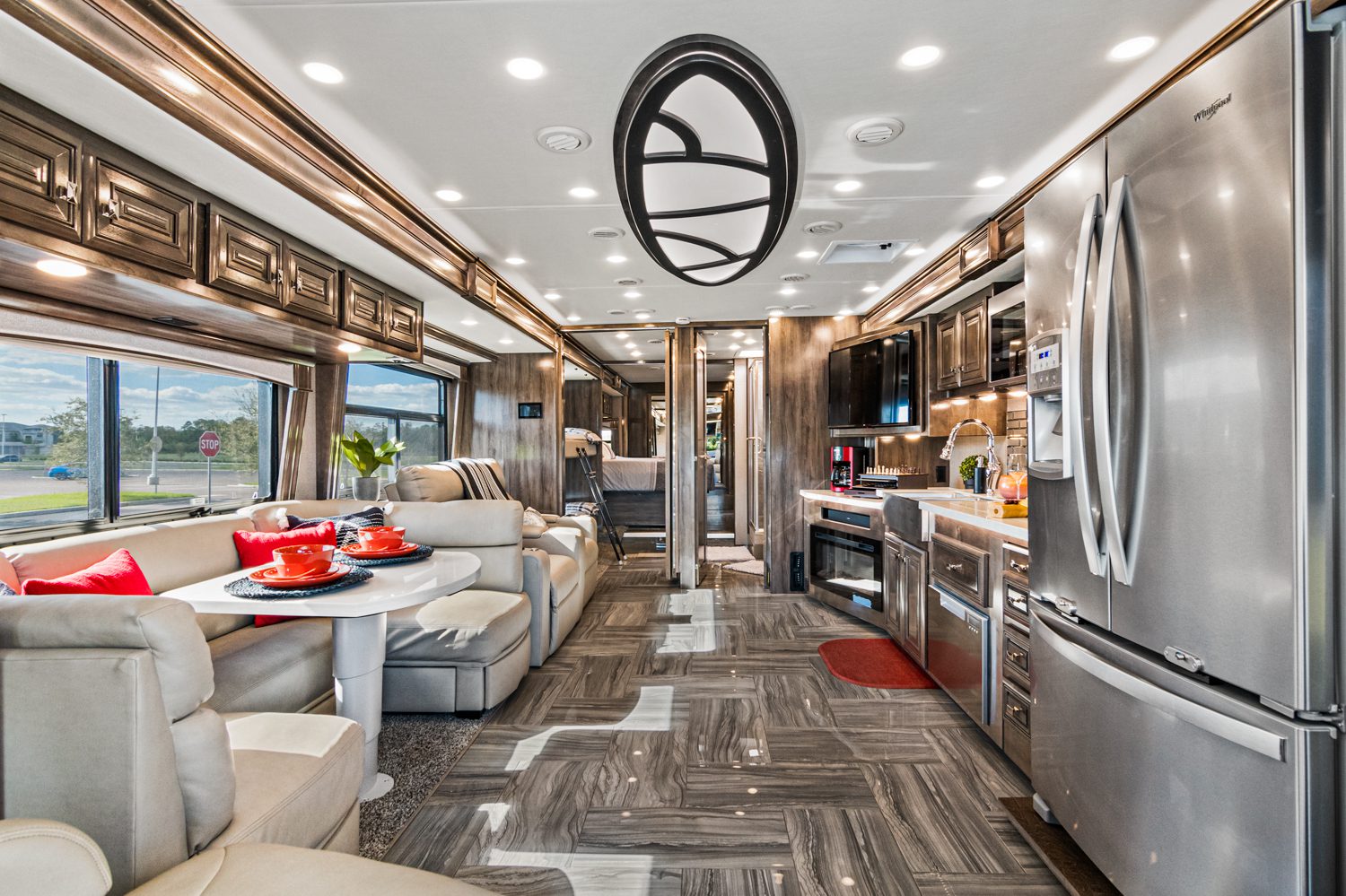Interior of a modern RV showing a kitchen area with stainless steel appliances, a dining nook, and a living space with sofas. The decor features overhead cabinets, tiled floors, and ceiling lighting.